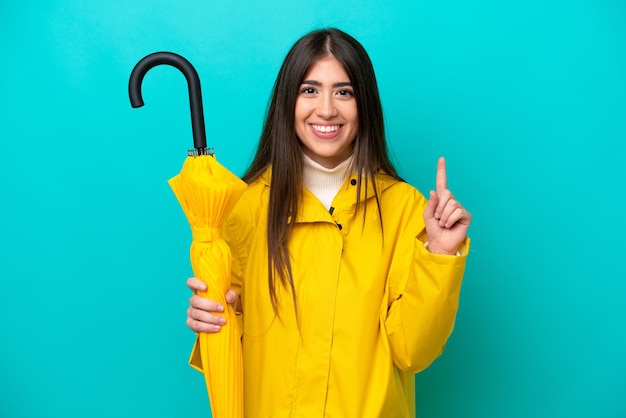 Young caucasian woman with rainproof coat and umbrella isolated on blue background pointing up a great idea