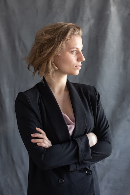 Young caucasian woman with short hair posing in black suit jacket in front of gray fabric