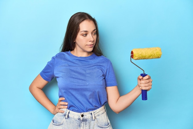 Young Caucasian woman with yellow paint roller on blue studio