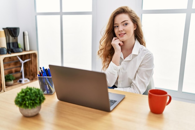 Young caucasian woman working at the office using computer laptop with hand on chin thinking about question pensive expression smiling and thoughtful face doubt concept