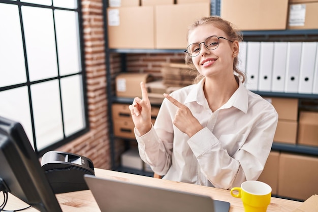 Young caucasian woman working at small business ecommerce using laptop smiling and looking at the camera pointing with two hands and fingers to the side