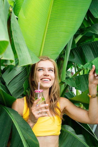 Young caucasian woman in yellow swuimsuit drinking beverage standing in tropical palm leaves