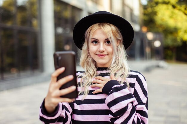 Young charismatic smiling woman in hat using phone outdoors