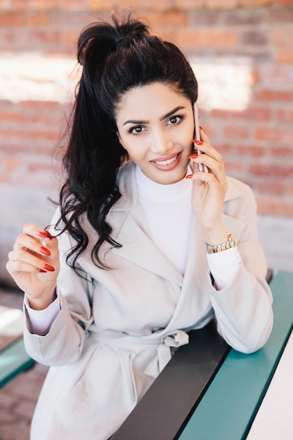 Young charismatic woman with dark wavy pony tail having red manicure dressed eleganltly talking on her mobile phone with smile while sitting at cafe waiting for her order Communication concept