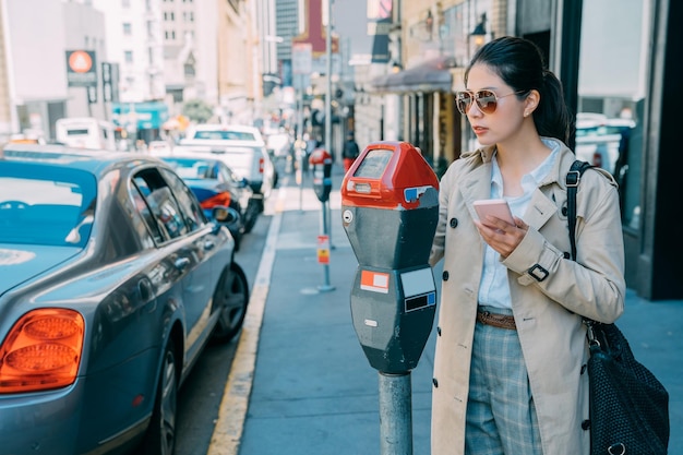 Young charming asian Japanese woman paying for car parking at machine with mobile phone online app. beautiful office lady local lifestyle in san francisco usa. female holding cellphone doing payment