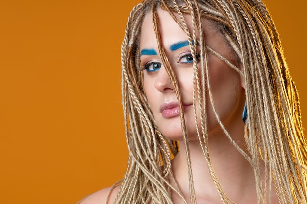 Young charming blonde with exotic braiding with microdreads, tinted eyebrows, blue eyebrows and makeup, photo on bright yellow