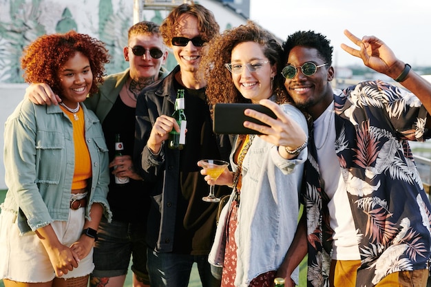 Young cheerfl woman with cocktail in martini glass making selfie with friends