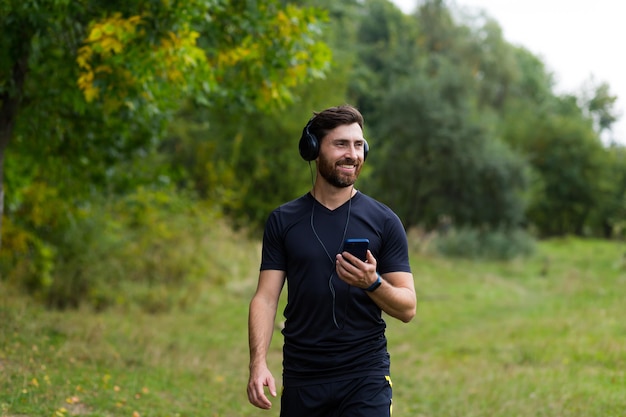 Young cheerful active guy sporty walking with headphones mobile phone enjoying listening to music, jogging outdoors on urban city park or forest background.