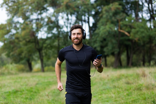 Young cheerful active guy sporty walking with headphones mobile phone enjoying listening to music, jogging outdoors on urban city park or forest background.