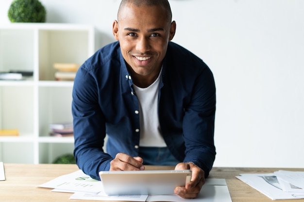 Young cheerful African man holding digital tablet and looking at camera with smile while standing at his working place