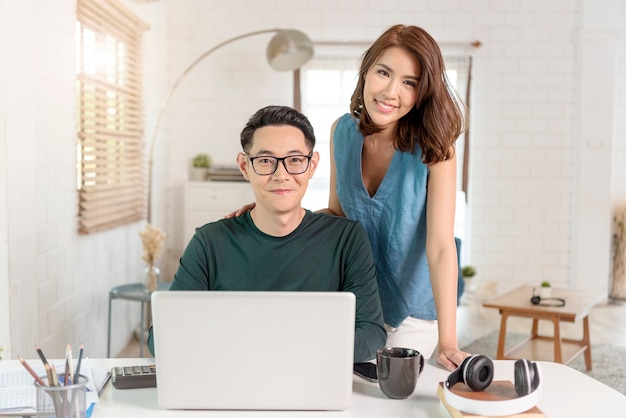 Young cheerful Asian couple colleagues work with laptop computer indoors in office talking with each other.