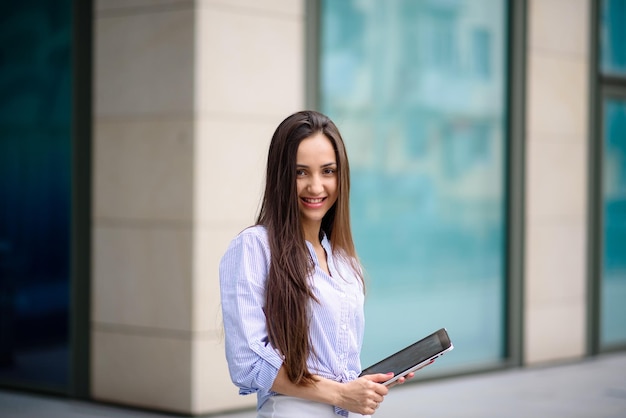 Young cheerful business lady in earphones looking at digital tablet with smile while standing on sunny street outdoors listening webinar or participating in online business meeting with team