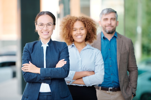 Young cheerful business leader in formalwear and her team behind looking at you with smiles while standing in row