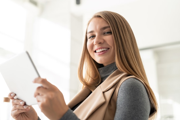 Young cheerful businesswoman with toothy smile looking at you while surfing in the net or working remotely in touchpad