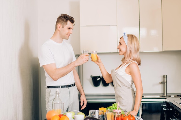 Young cheerful caucasian couple starting a new day with energy drink. Photo of relaxed happy young people holding glasses with orange juice and looking at each other with smile.