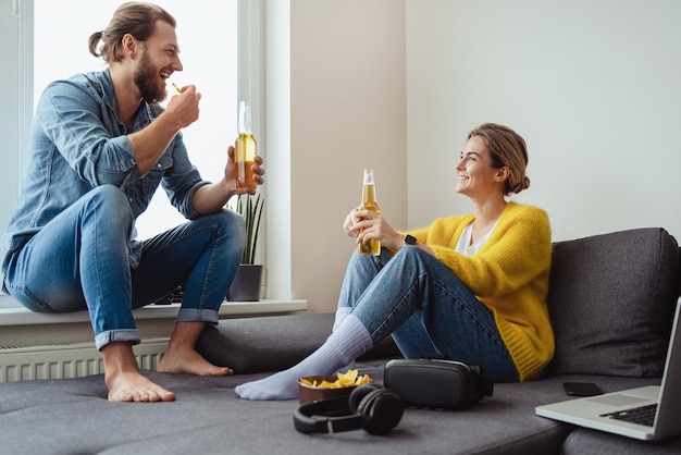 Young cheerful couple sitting on the sofa drinking beer and eating nachos