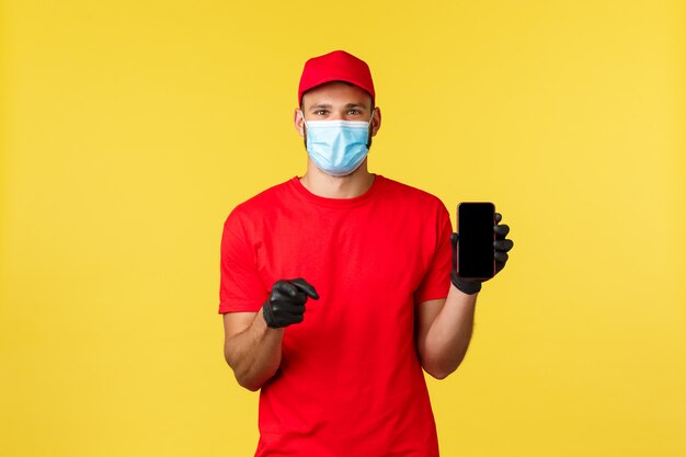 Young cheerful courier in medical mask and gloves, red uniform, show smartphone app, screen phone and pointing camera