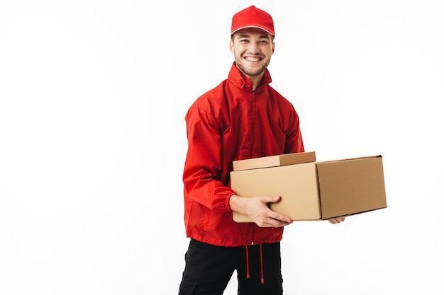 Young cheerful delivery man in red cap and jacket holding boxes in hands happily 