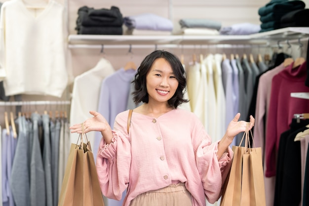 Young cheerful female shopaholic in smart casualwear holding two packs of big paperbags with purchases while standing in clothing department