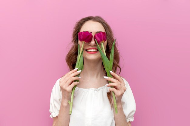 Young cheerful girl closes her eyes with two pink tulippans and smiles on pink isolated background