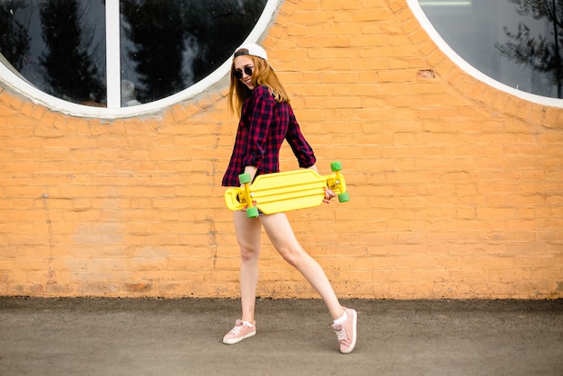 Young cheerful girl posing with yellow skateboard against orange wall.