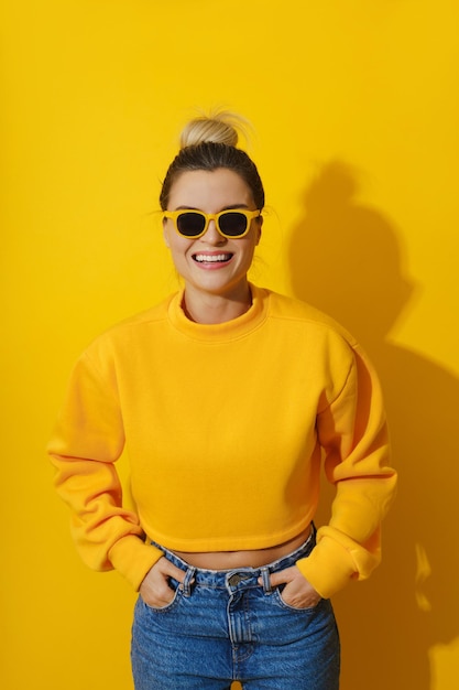 Young cheerful girl wearing yellow sweatshirt and sunglasses against yellow background