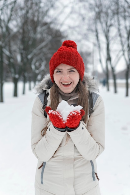 Young cheerful girl in winter park holds snow on her palms. Portrait of woman in knitted red hat.