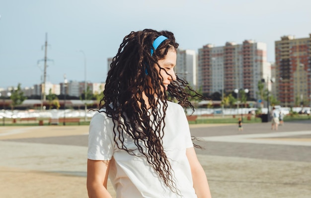 A young cheerful happy woman with dreadlocks dressed in a white Tshirt dancing listening to music with headphones resting relaxing in a city park walking along an alley Urban lifestyle concept