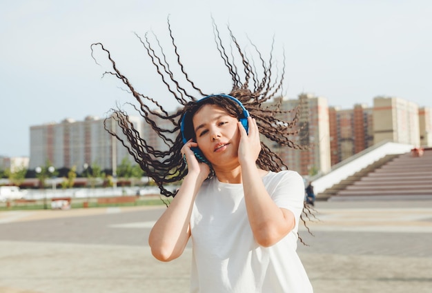 A young cheerful happy woman with dreadlocks dressed in a white Tshirt dancing listening to music with headphones resting relaxing in a city park walking along an alley Urban lifestyle concept