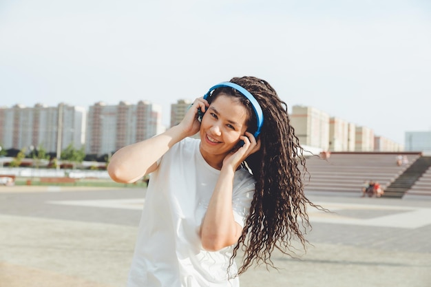 A young cheerful happy woman with dreadlocks dressed in a white Tshirt dancing listening to music with headphones resting relaxing in a city park walking along an alley Urban lifestyle concept