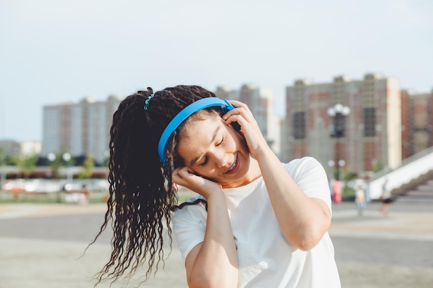 A young cheerful happy woman with dreadlocks dressed in a white Tshirt dancing listening to music with headphones resting relaxing in a city park walking along an alley Urban lifestyle concept