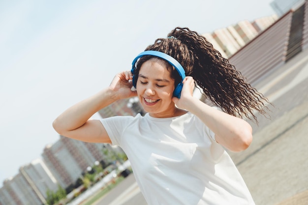 A young cheerful happy woman with dreadlocks dressed in a white Tshirt dancing listening to music with headphones resting relaxing in a city park walking along an alley Urban lifestyle concept