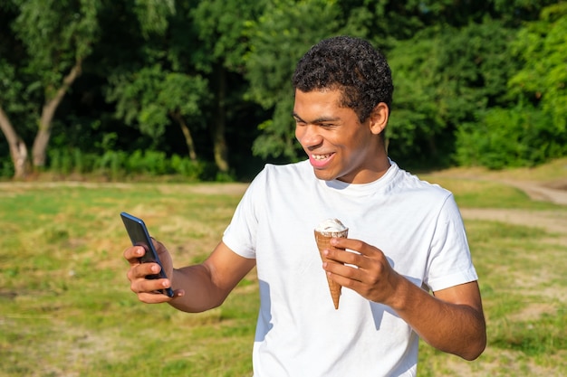 Young cheerful and laughing African-American and caucasian man eats ice cream and use smartphone, makes selfie while standing in park outdoors in summer