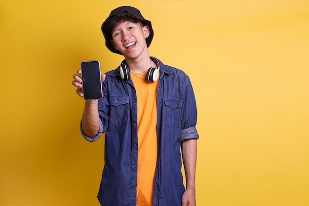 Young cheerful male student in casual clothes bucket hat with headphones on his neck showing blank