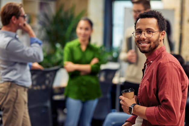 Young cheerful mixed race man holding paper coffee cup and smiling at camera while working