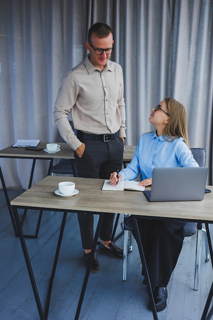 Young cheerful multinational corporate lawyers in formal wear editing text of new contract using laptop while sitting at table with notepad in modern office