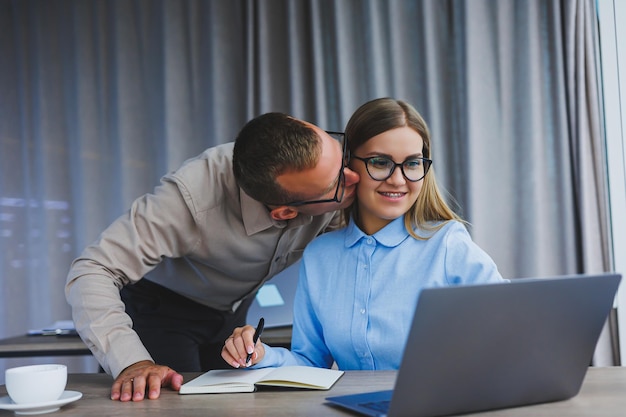 Young cheerful multinational corporate lawyers in formal wear editing text of new contract using laptop while sitting at table with notepad in modern office