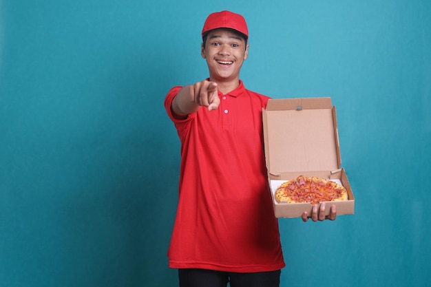 Young cheerful pizza man in red t-shirt and cap holding an open pizza box pointing at you