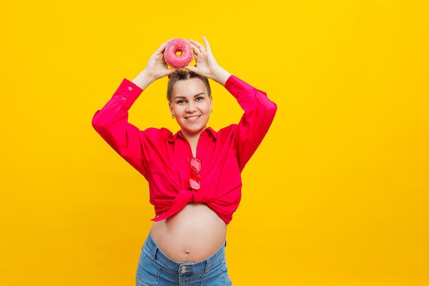 Young cheerful pregnant woman in pink shirt isolated on yellow background holding donuts with a cheerful expression on her face Sweet food during pregnancy