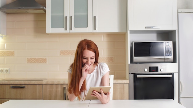 Young cheerful redhead girl sitting at the table in the kitchen reading the good news on her tablet