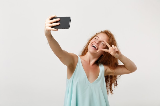 Young cheerful redhead woman girl in casual light clothes posing isolated on white background. People lifestyle concept. Mock up copy space. Doing selfie shot on mobile phone, showing victory sign.