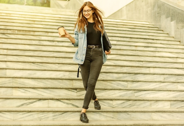 Young cheerful student woman in a denim jacket and glasses goes down the stairs with a laptop and a coffee cup in her hands in the city