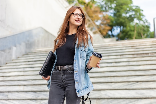 Young cheerful student woman in a denim jacket and glasses goes down the stairs with a laptop and a coffee cup in her hands in the city