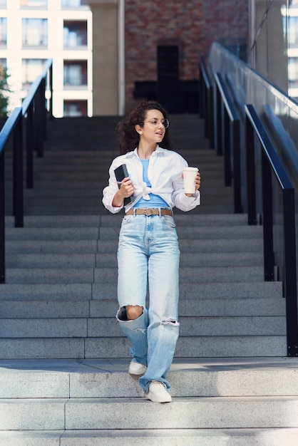 Young cheerful student woman in glasses goes down the stairs with cup of coffee and notebook in her