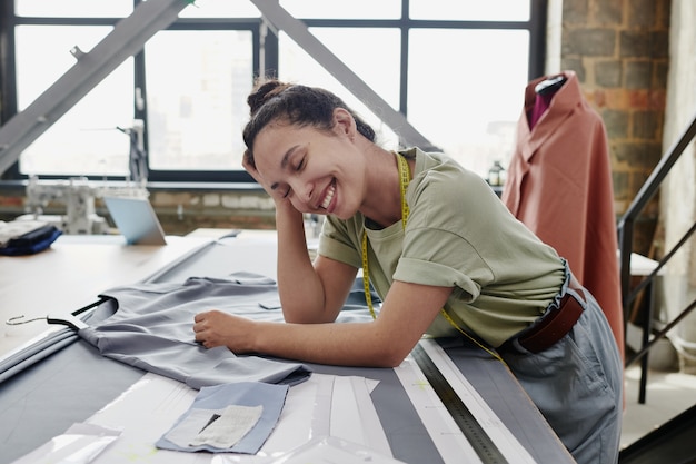 Young cheerful tailor or designer of clothes in casualwear leaning on table with unfinished dress, textile, paper patterns and ruler during work