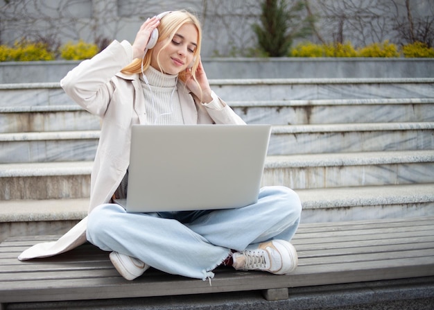 Young cheerful woman listening to music in headphones while sitting on a bench with a laptop