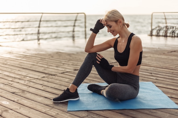 Young cheerful woman in sportswear sits on a mat on the beach at sunrise