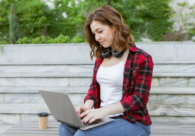 Young cheerful woman student is using laptop outdoors. Distance work or education concept