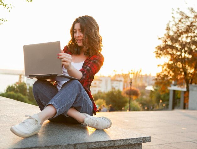 Young cheerful woman student sitting with laptop outdoors early in the morning at sunrise