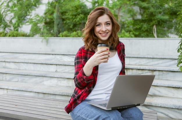 Young cheerful woman student using laptop and drink coffee outdoors. Distance work or education concept